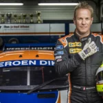 Jeroen Dik, a Dutch diesel racing champion, posing in front of his race car in a racing suit, holding his helmet with trophies visible in the background.