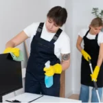Two professional cleaners in black aprons and yellow gloves performing deep cleaning — one wiping a monitor screen with a spray bottle and cloth, the other mopping the floor in a bright indoor space.